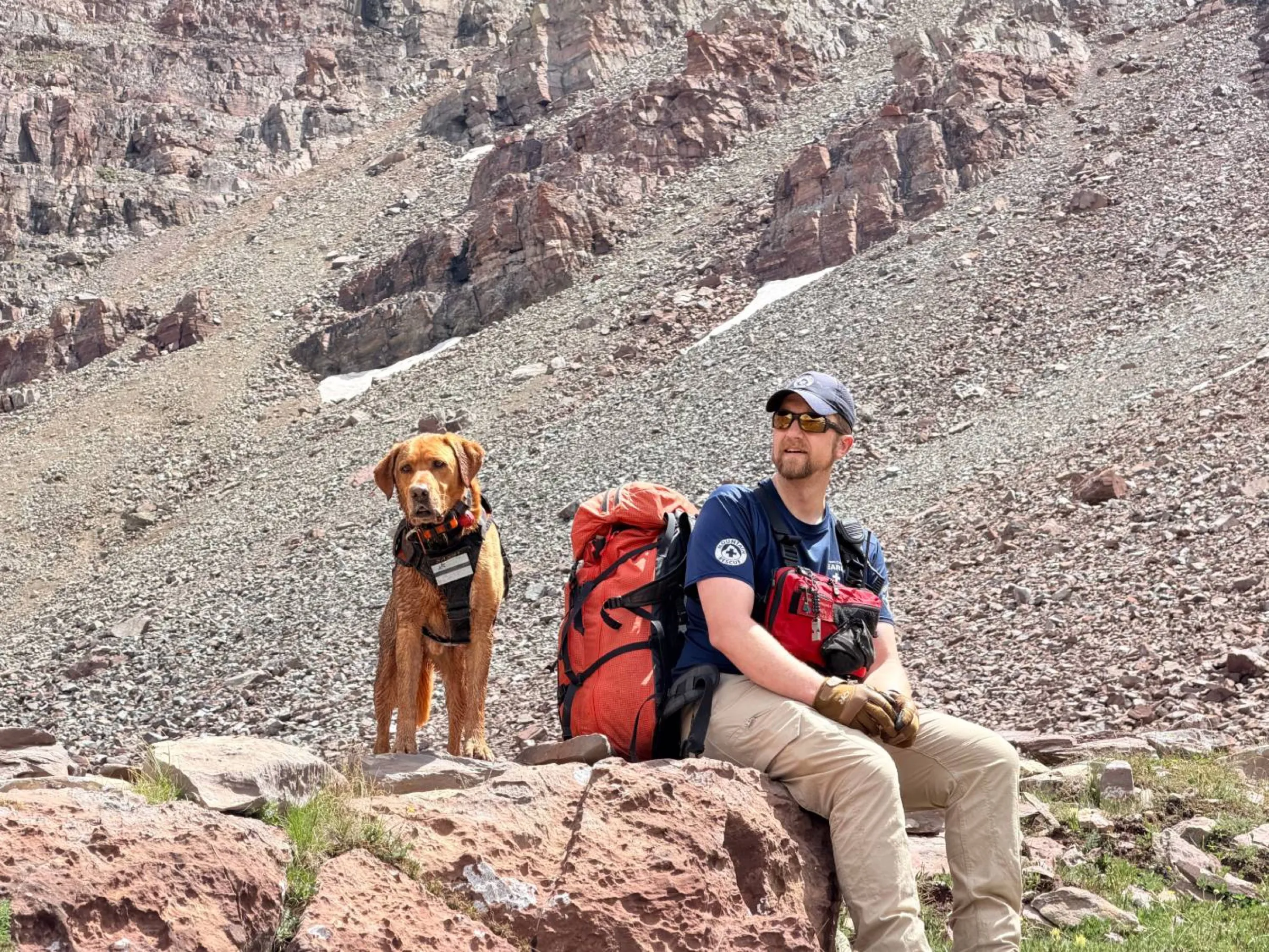 Jay Christianson in a search and rescue shirt, sitting on a rock beside an orange pack, with Saxby, a red-coated dog in a working harness, standing on the talus behind, below red cliffs.