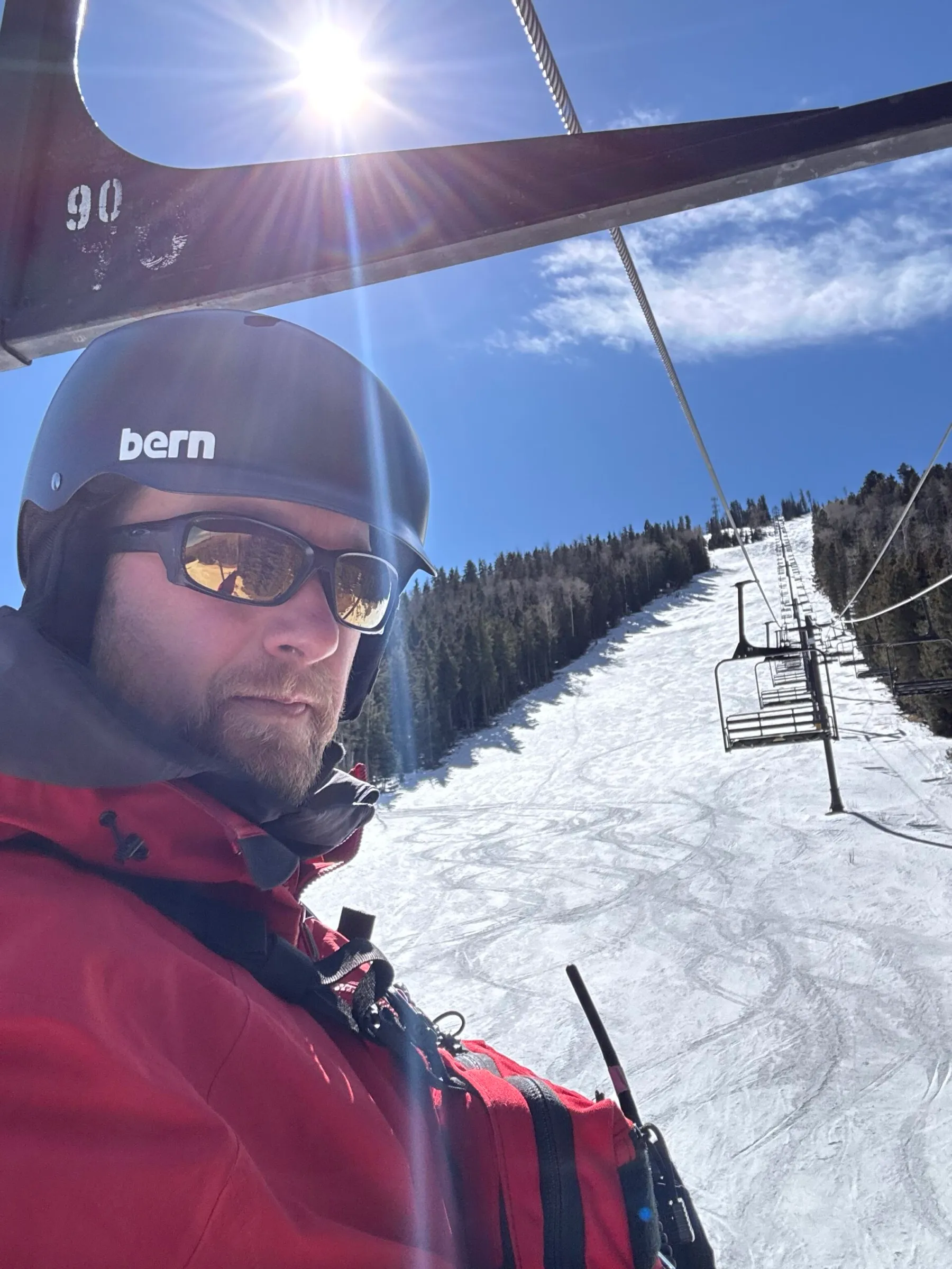 Jay Christianson in a helmet and red jacket on a chairlift, with a ski run below.